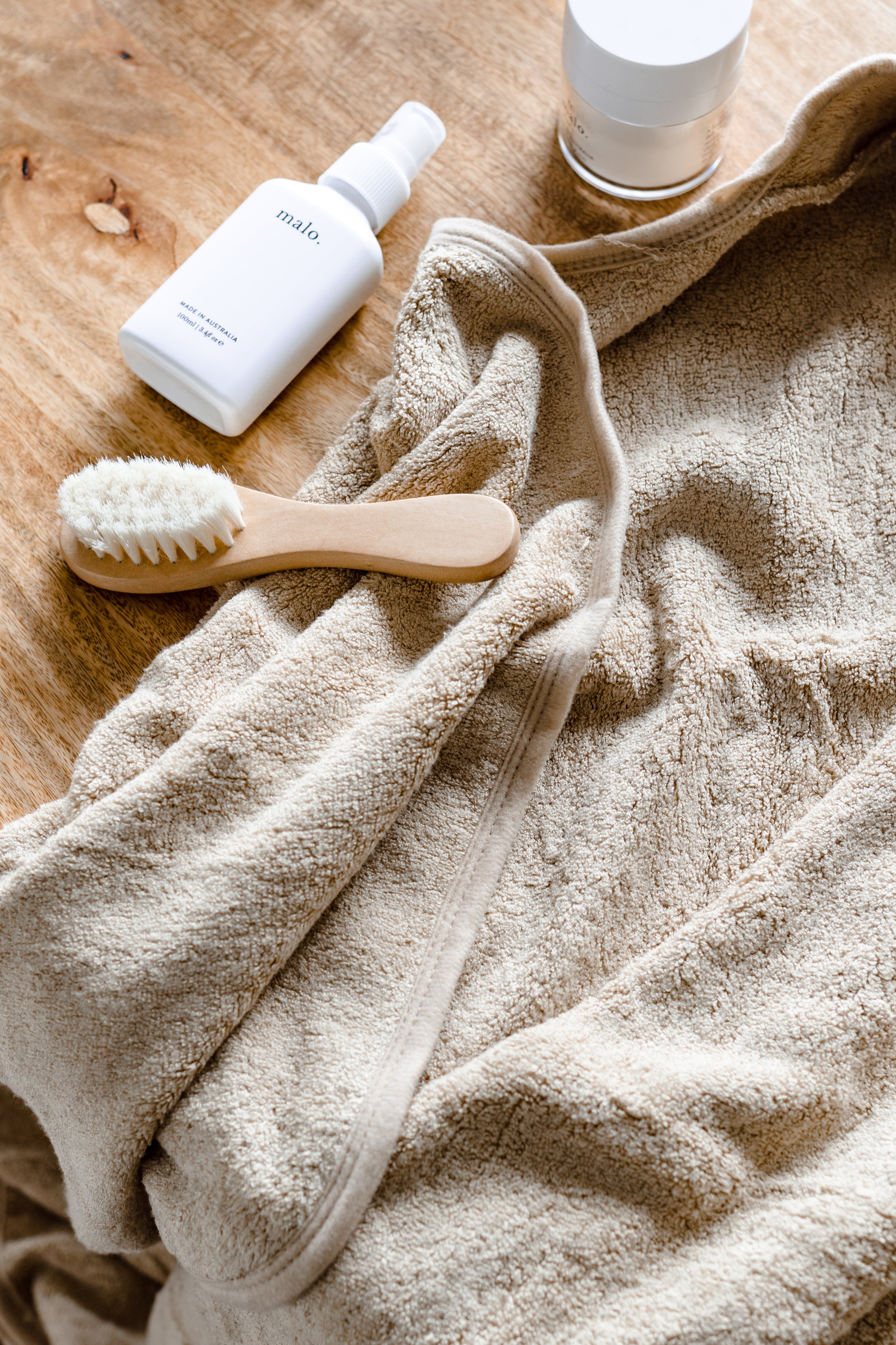 A beige towel, a wooden brush, a white bottle labeled "malo," and a small white jar are laid out on a wooden surface.