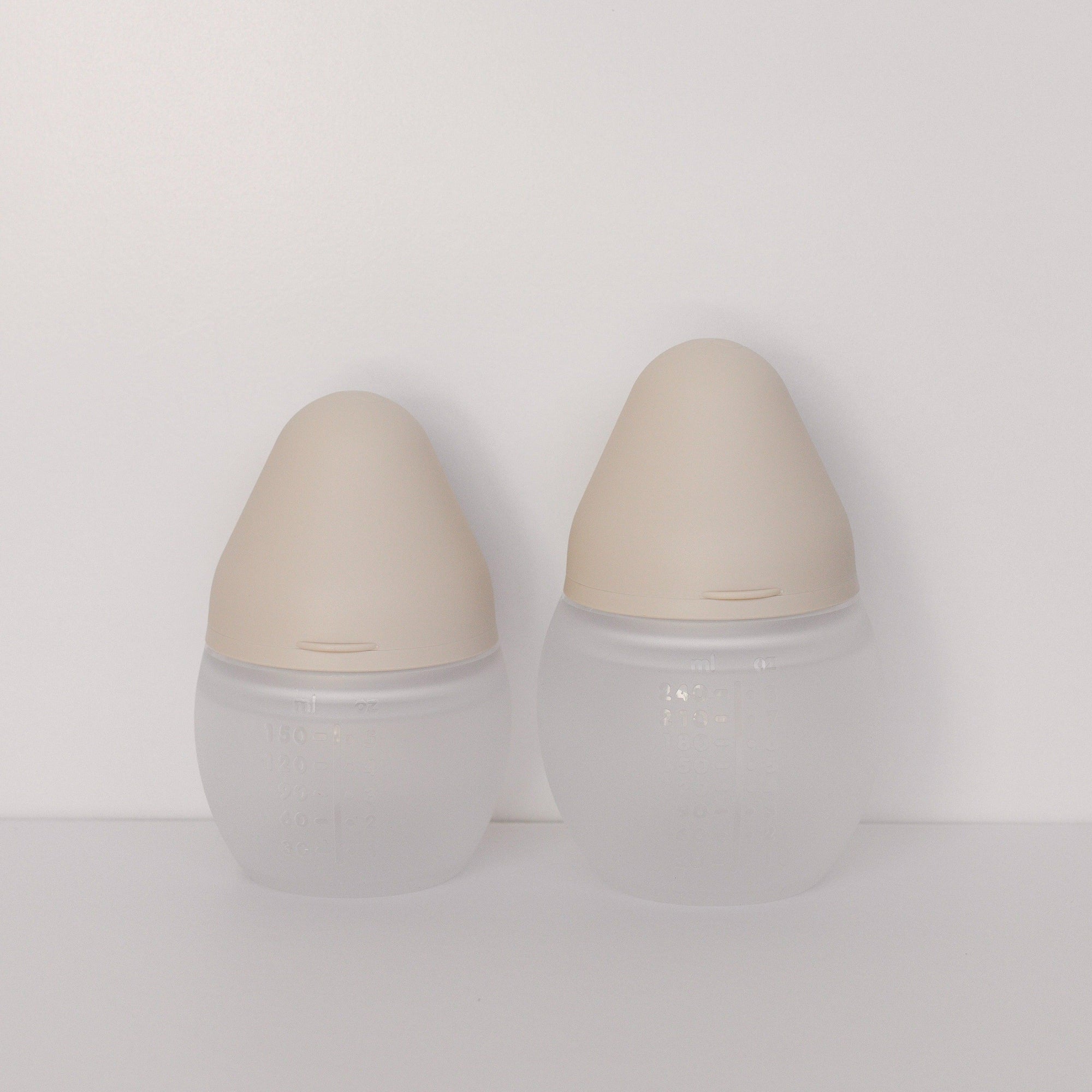 Two Elhée France baby bottles in the shade sand standing against a white surface.